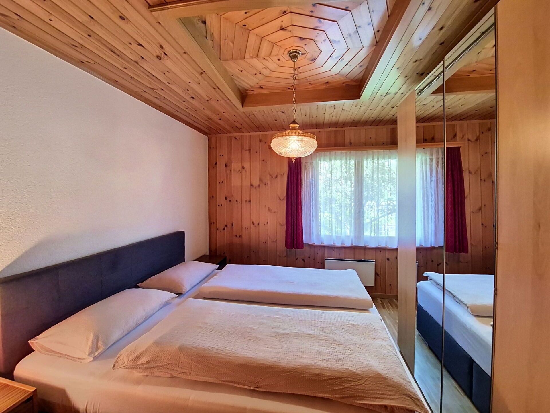 Ein Zimmer mit Holzwänden und -decke, einem Bett mit weißer Bettwäsche, einer Hängelampe und einem Fenster mit Vorhängen.
