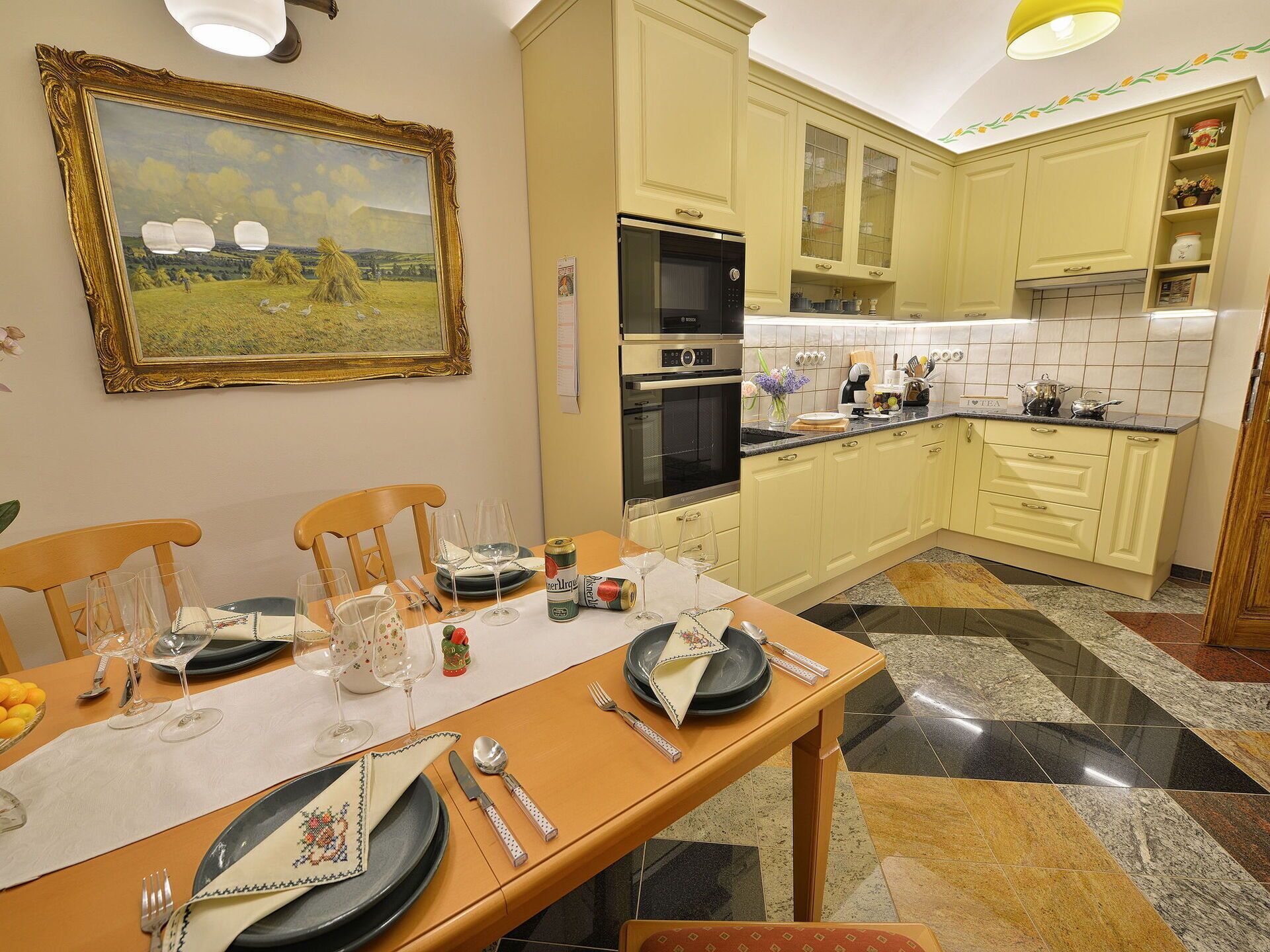 Uma cozinha bem iluminada com uma mesa de jantar posta para uma refeição, um forno Bosch e um quadro de uma paisagem na parede.