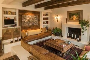 A living room with a brown leather sofa, a coffee table with books, a fireplace, and a wall-mounted TV. - Spacious spectacular 9-bedroom Fresnaye Villa (Cape Town)