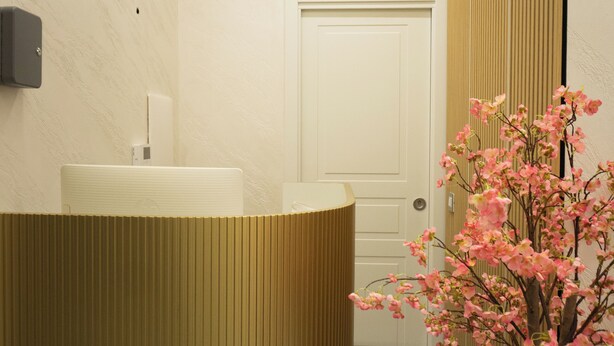 Un área de recepción con un mostrador curvo, un teléfono en la pared, una puerta y una planta en maceta con flores rosadas.