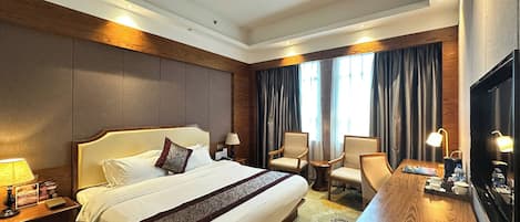 Quarto de hotel com uma cama grande, duas poltronas, uma mesinha, televisão e janela com cortinas.