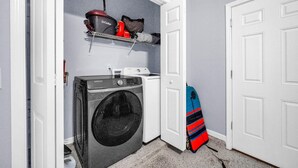 A laundry room with a washing machine, a dryer, a shelf with a vacuum cleaner, and a surfboard. - Shaker of Salt (Panama City Beach)