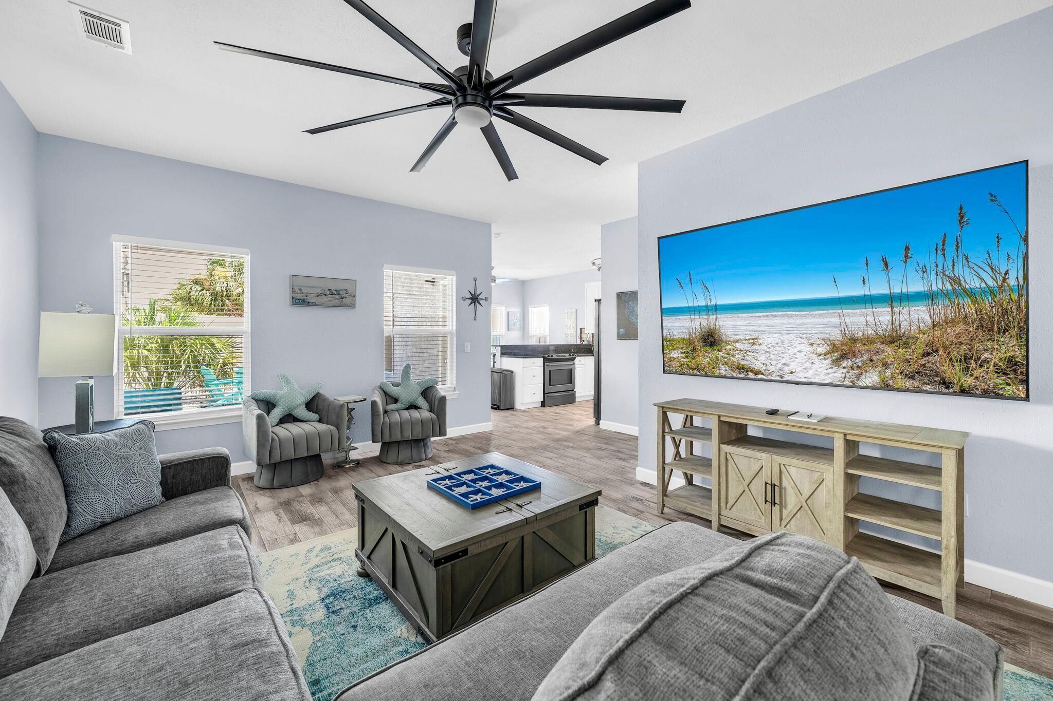 Een moderne woonkamer met een plafondventilator, een grote flatscreen televisie, grijze banken, een salontafel en uitzicht op het strand door een raam.