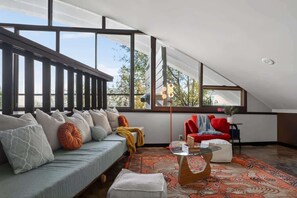A modern loft-style room with a sloped ceiling, large windows, a sofa with cushions, a red chair, and a patterned rug. - Mid-Century Mountain View Getaway for 4 (Billings)