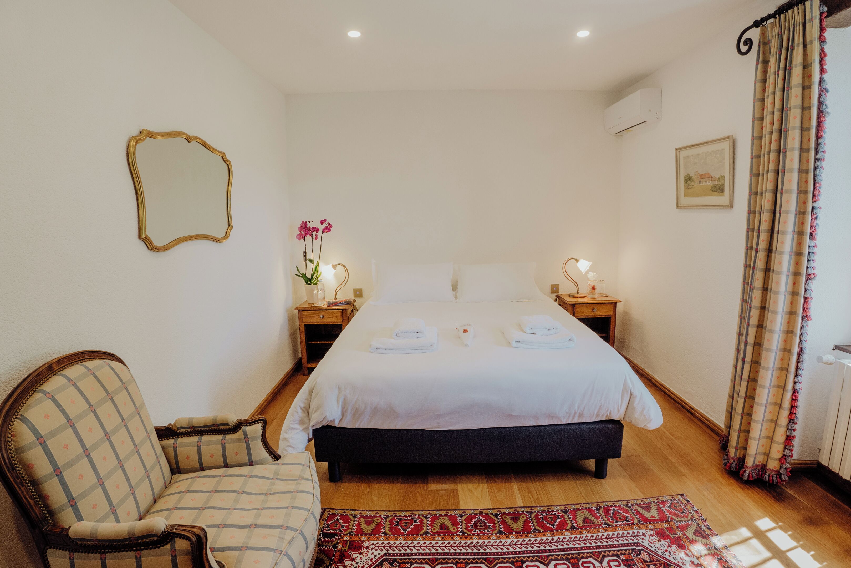 Une chambre à coucher comprenant un lit, une chaise, une table de chevet avec une lampe, un miroir et une fenêtre avec des rideaux.