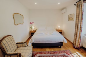 A bedroom with a bed, a chair, a bedside table with a lamp, a mirror, and a window with curtains.