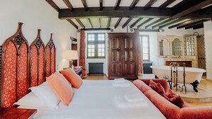 Quarto com uma cama grande, um sofá vermelho estofado, um guarda-roupa de madeira, uma banheira de patas de leão e um espelho com painel de azulejos.