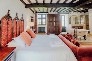 Une chambre avec un grand lit, un canapé rouge recouvert de tissu, une armoire en bois, une baignoire sur pieds et un miroir avec un pare-éclaboussures carrelé.