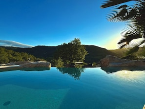 A clear view of a swimming pool with a mountain backdrop, surrounded by rocks and greenery. - Château de Tournou – Sleeps 15, 7 Bedrooms & Private Infinity Pool (Castelreng)