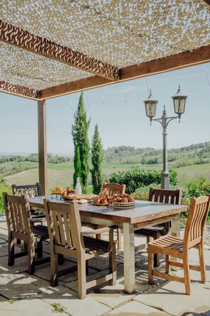 Un espacio de comedor al aire libre con una pérgola, sillas de madera, una mesa con pan y pasteles, y vistas a colinas ondulantes.