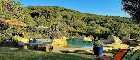 Zona de piscina con sillones de descanso, una sombrilla de paja y montañas al fondo.