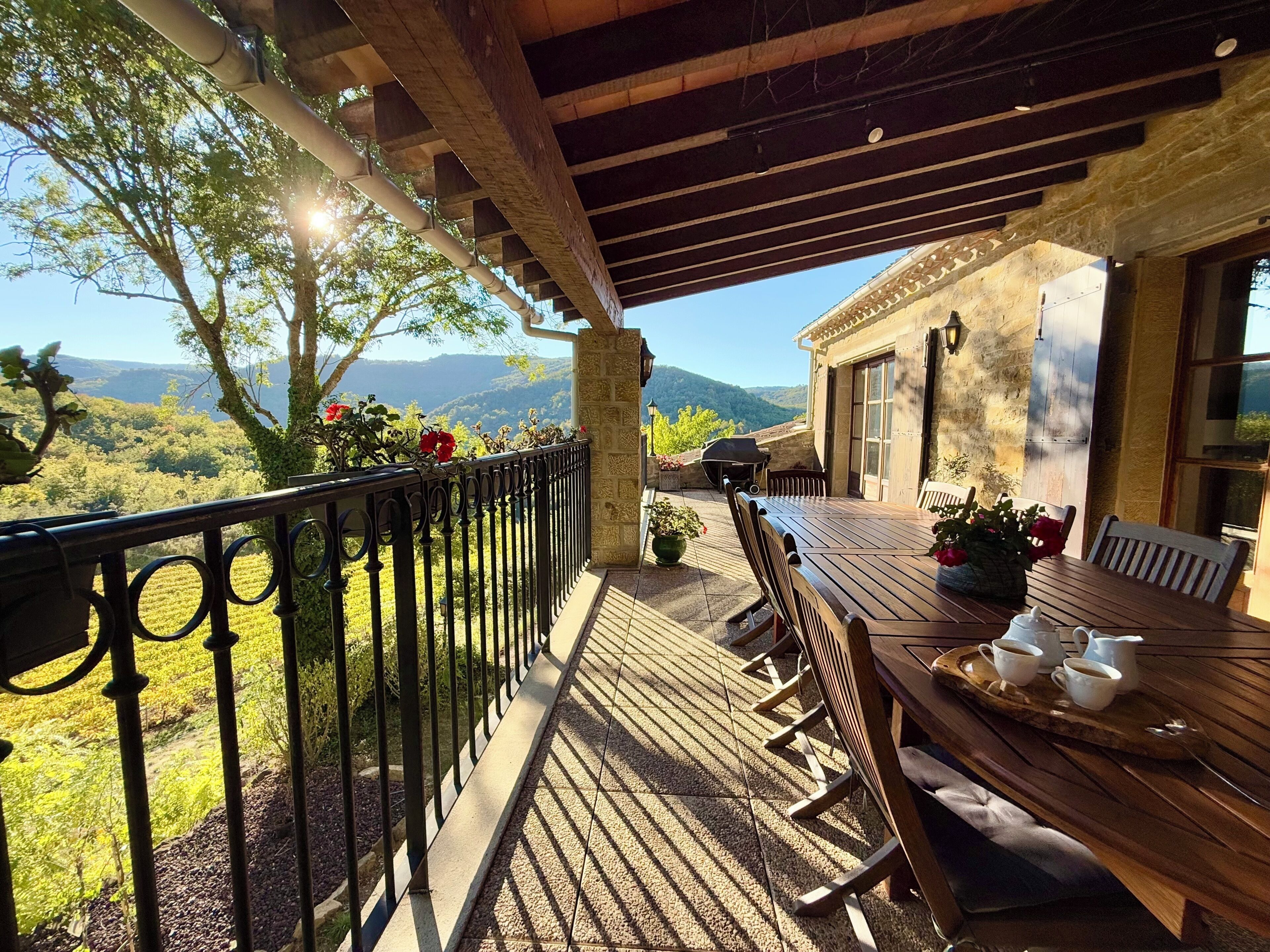A covered outdoor patio with a wooden table and chairs, a stone wall, and a view of the countryside.