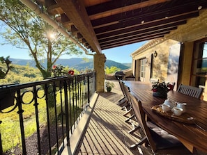 A covered outdoor patio with a wooden table and chairs, a stone wall, and a view of the countryside. - Château de Tournou – Sleeps 15, 7 Bedrooms & Private Infinity Pool (Castelreng)