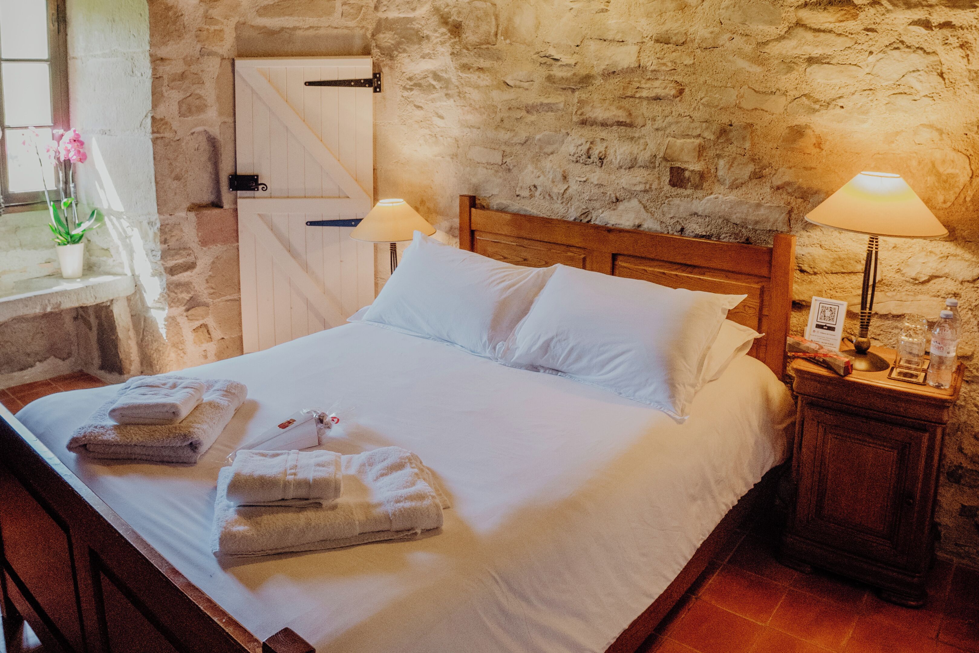 Un lit bien fait, avec du linge de lit et des serviettes blanches, une tête de lit en bois, un mur en pierre et une table de chevet avec une lampe et une petite table.