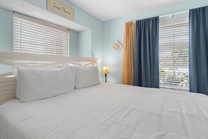 Une chambre à coucher avec un lit, du linge de lit blanc, une table de chevet avec une lampe et une fenêtre avec des stores et des rideaux.