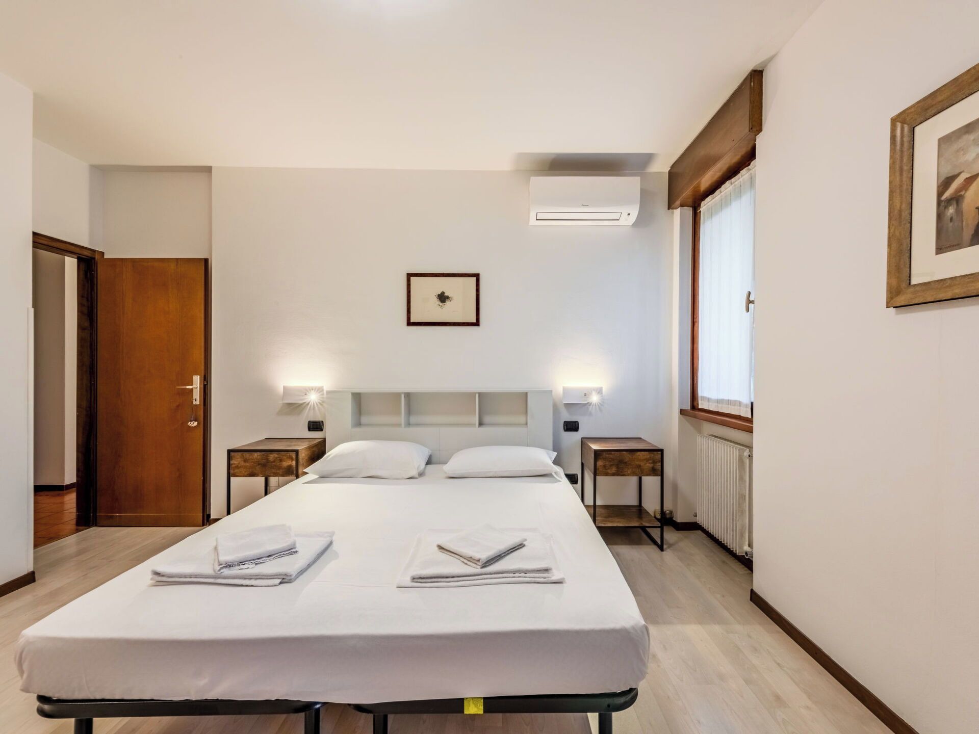 A hotel room with a bed, bedside tables, a framed picture, an air conditioning unit, and a window.