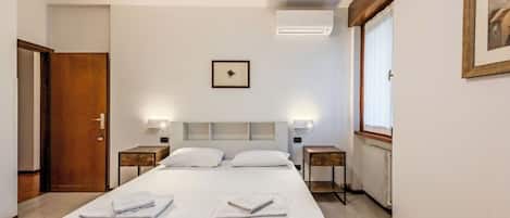 A hotel room with a bed, bedside tables, a framed picture, an air conditioning unit, and a window.