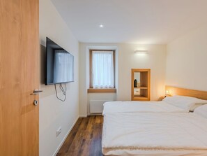 Quarto de hotel com cama, televisão na parede, janela com cortinas, mesa de cabeceira com abajur e porta de madeira.