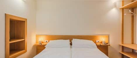 A hotel room with a double bed, bedside tables with lamps, a wooden shelf unit, and a hanging clothes rack.