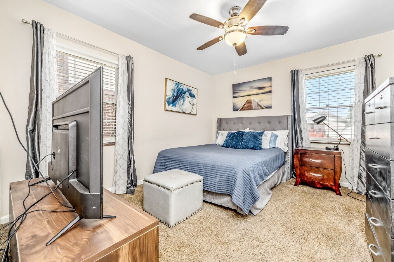 A bedroom with a bed, a desk, a dresser, a ceiling fan, and artwork on the walls.