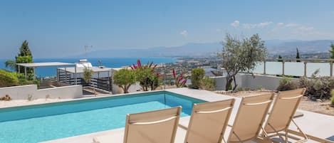 A pool area with lounge chairs and a view of the sea and mountains.