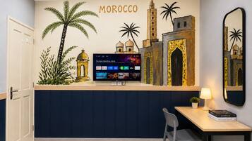 A room with a wall mural depicting Moroccan architecture, a flat-screen TV mounted on a wall, a desk with a chair, and a lamp.