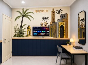Habitación con un mural que representa arquitectura marroquí, un televisor de pantalla plana en la pared, un escritorio con una silla y una lámpara.