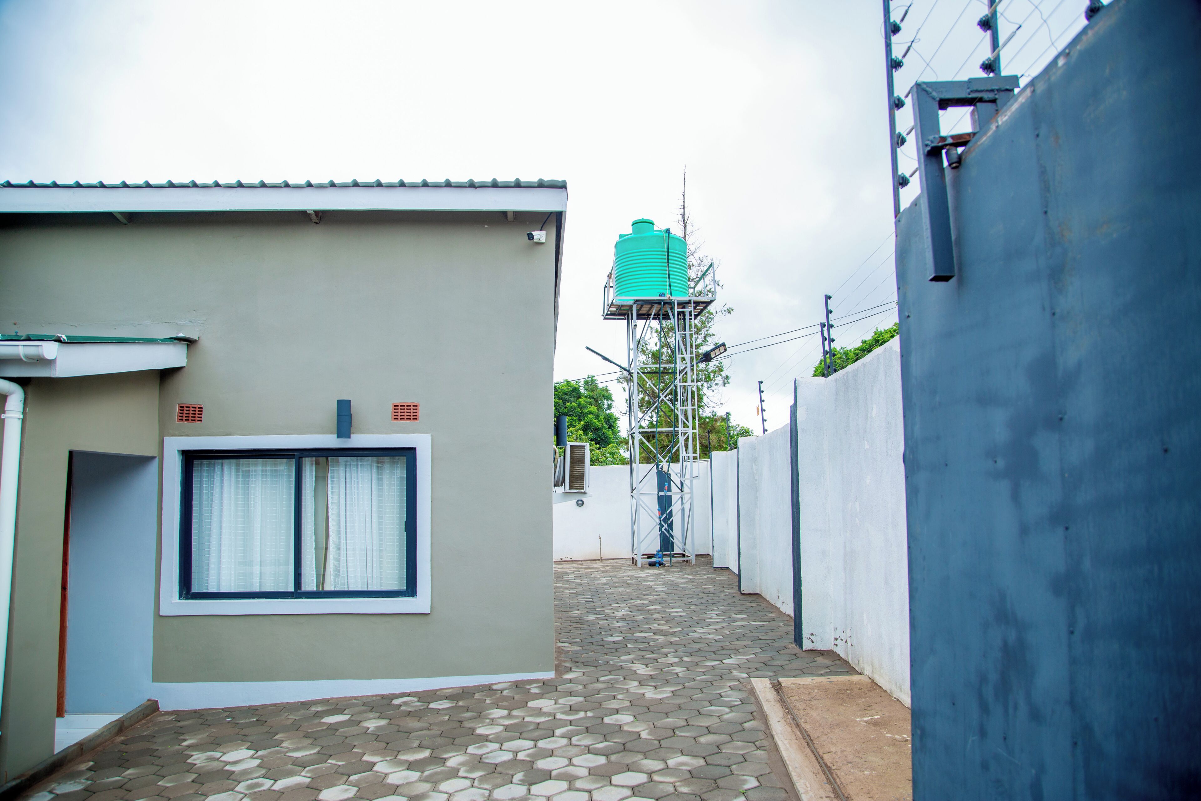 A narrow alleyway with a building on the left, a water tank on a metal structure in the distance, and a blue wall on the right.