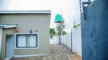 A narrow alleyway with a building on the left, a water tank on a metal structure in the distance, and a blue wall on the right.