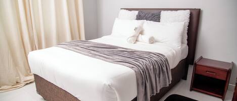 A neatly made bed with white linens and a grey blanket, a wooden nightstand with a drawer, and a black rug on a tiled floor.