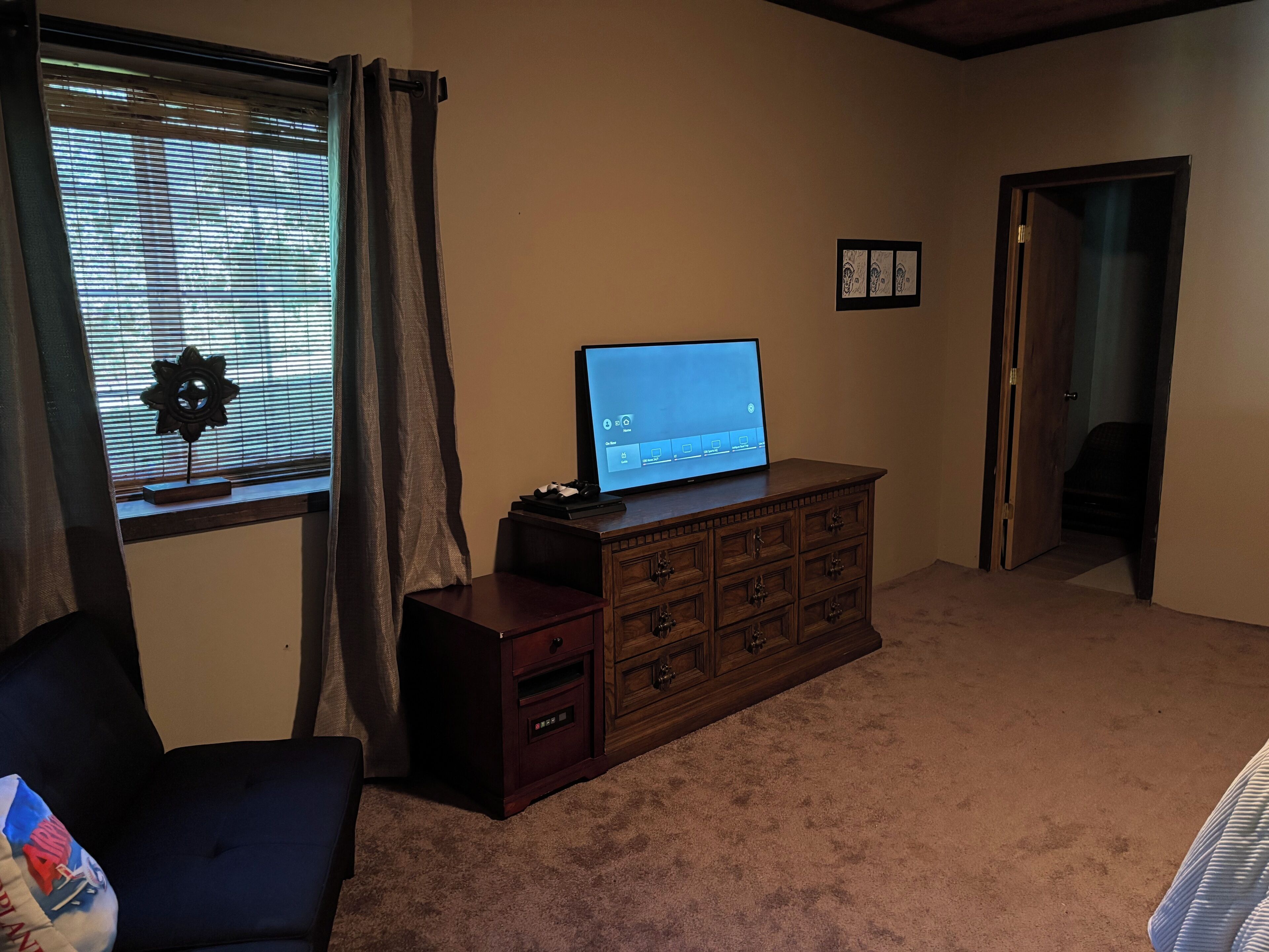 Una habitación con un aparador de madera, un televisor encima, una ventana con persianas, una puerta y piso alfombrado.