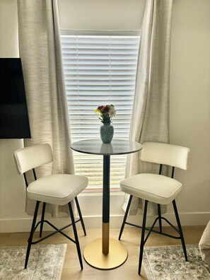 Un comedor con una mesa redonda, dos sillas, un florero con flores y una ventana con persianas.