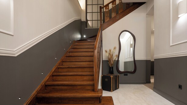 A wooden staircase with a dark wall on one side and a light wall on the other, leading to a landing with a mirror and a vase. - Kosmitis Suites Athens (Athens)