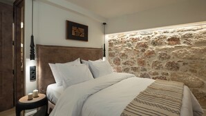 A bedroom with a stone wall, a bed with white bedding, a small bedside table with candles, and a wall-mounted light fixture. - Kosmitis Suites Athens (Athens)