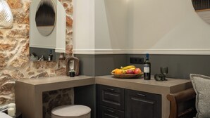 A corner kitchen with a stone wall, a fruit bowl, wine bottle, and a glass. - Kosmitis Suites Athens (Athens)
