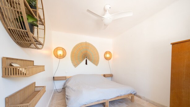 Een slaapkamer met een bed, houten hoofdbord, wandplanken, een plafondventilator en een lamp in de vorm van een ventilator.