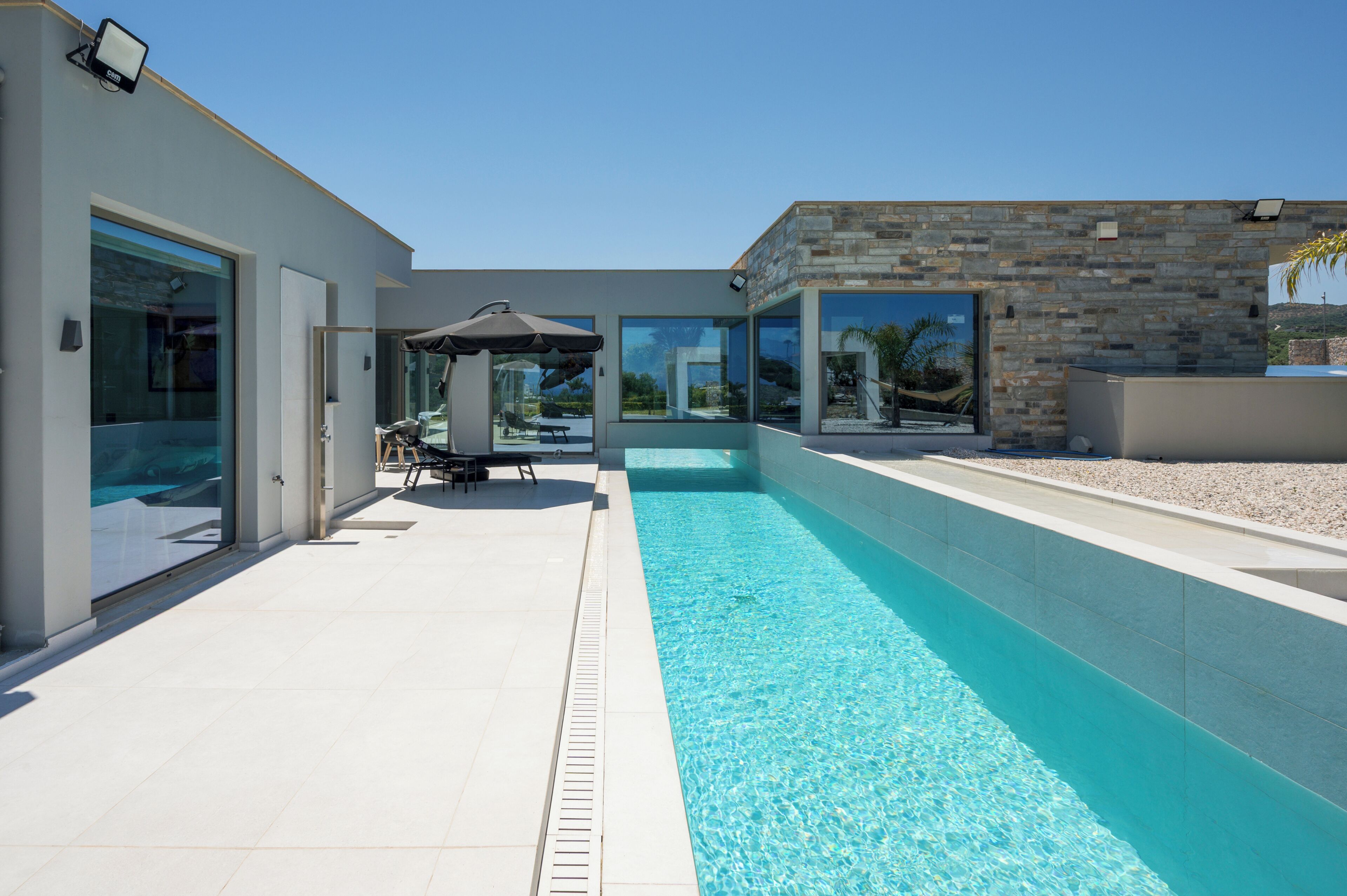 A modern outdoor pool area with a clear blue pool, a patio with a table and chairs, and a stone wall.