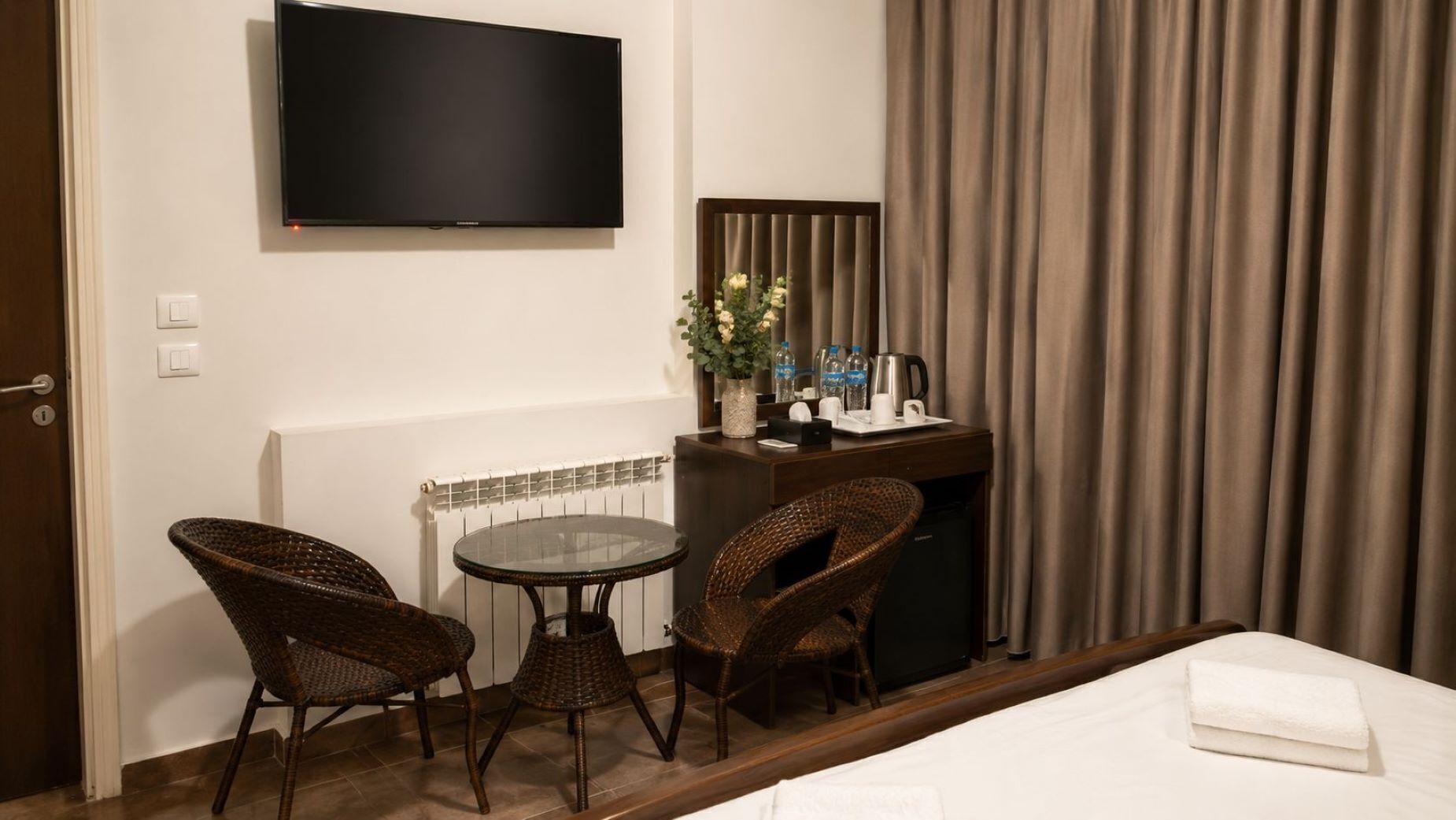 Quarto de hotel com cama, escrivaninha com cadeira, televisão, mesinha e janela com cortinas.