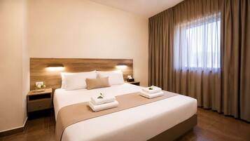A hotel room with a large bed, wooden headboard, bedside tables, a nightstand, a window with curtains, and a wall-mounted light.