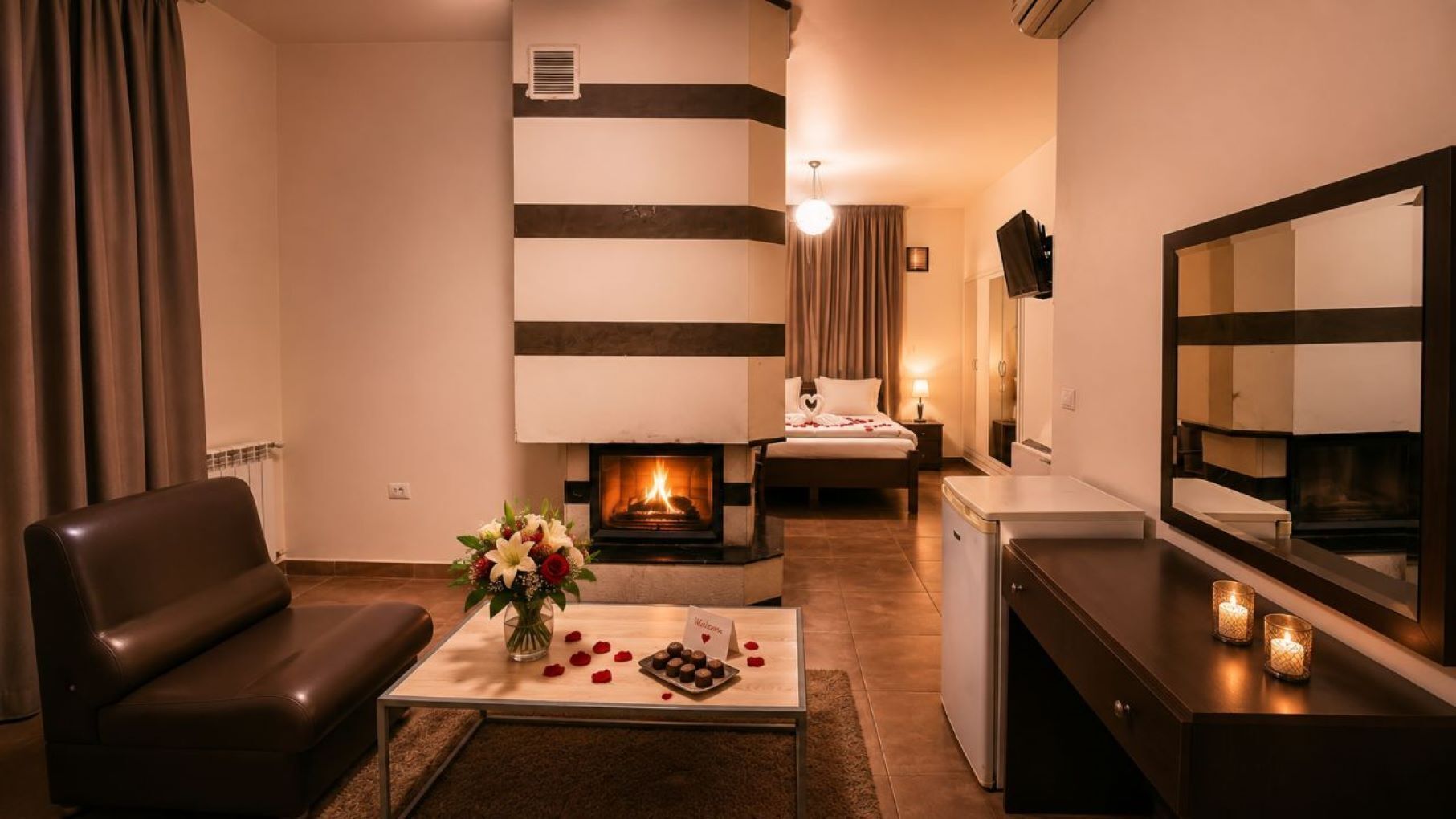 A cozy hotel room with a fireplace, a bed, a sitting area with a leather chair, and a small table with a welcome card and flowers.
