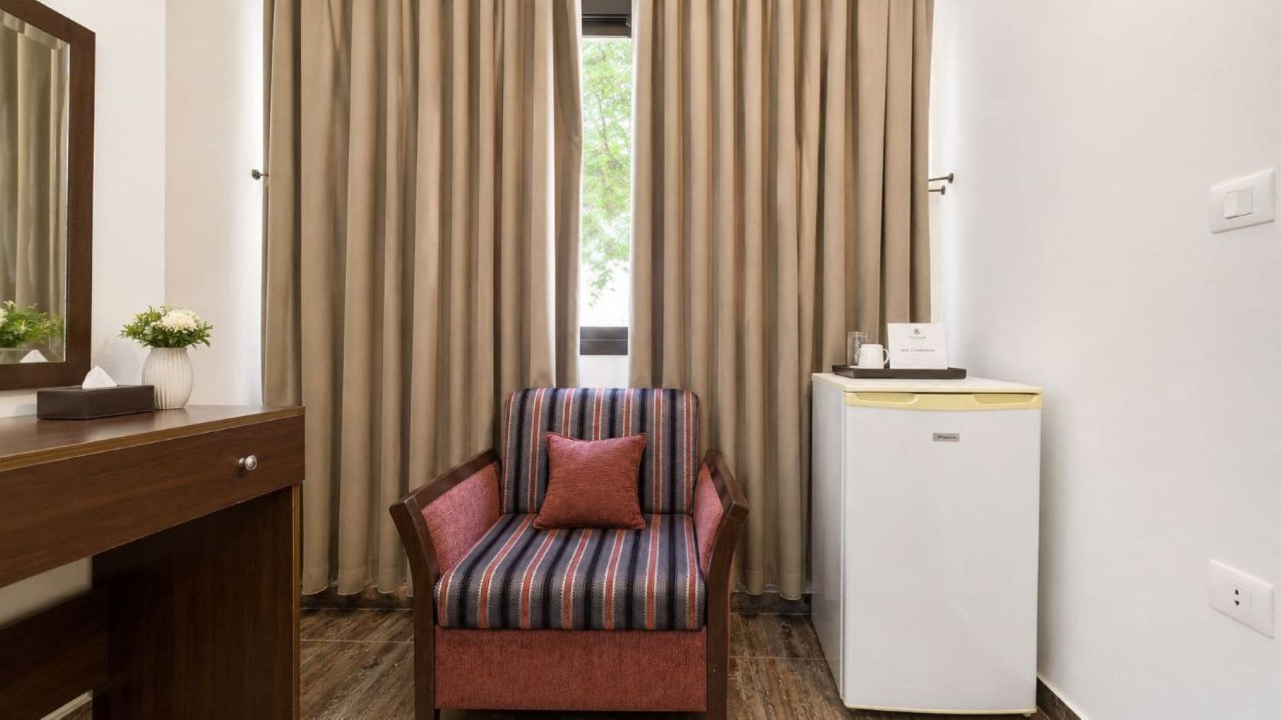 A hotel room with a striped armchair, a small table with a cup and a menu, and a compact refrigerator.