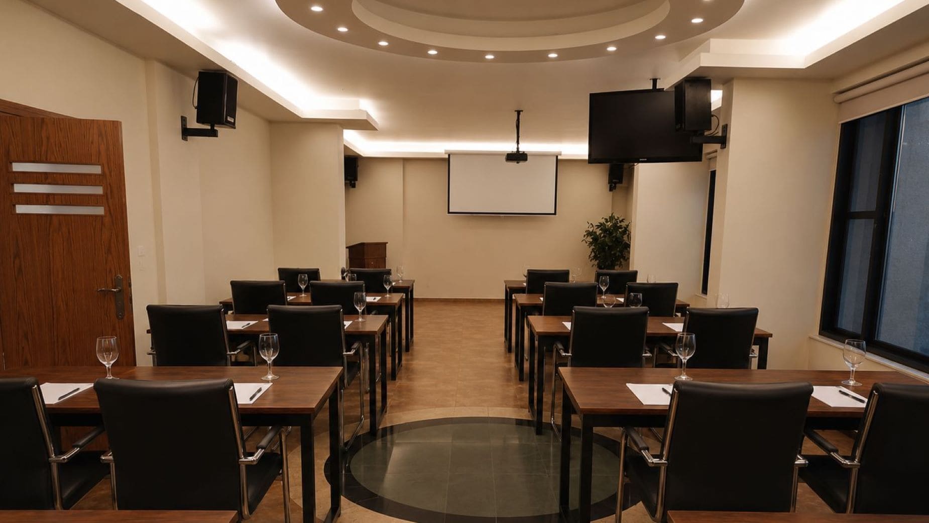 Una sala de conferencias con una pantalla grande, varias mesas con sillas y una puerta de madera.