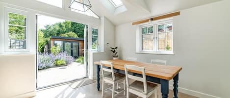 A dining area with a wooden table and chairs, large windows, and a view of the garden.