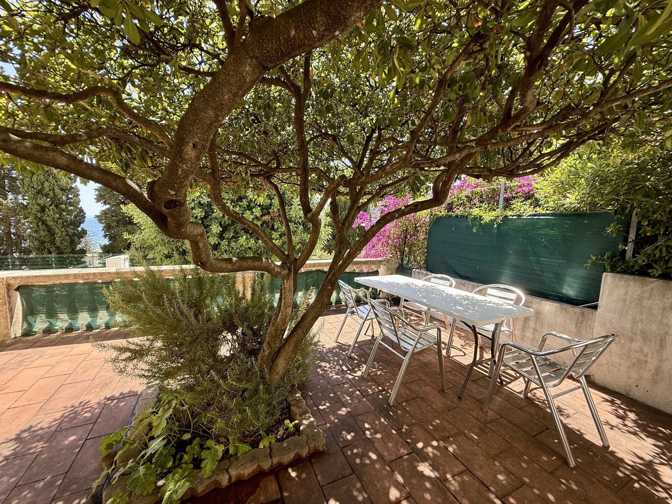 A patio with a table and chairs, surrounded by greenery and a view of the sea.