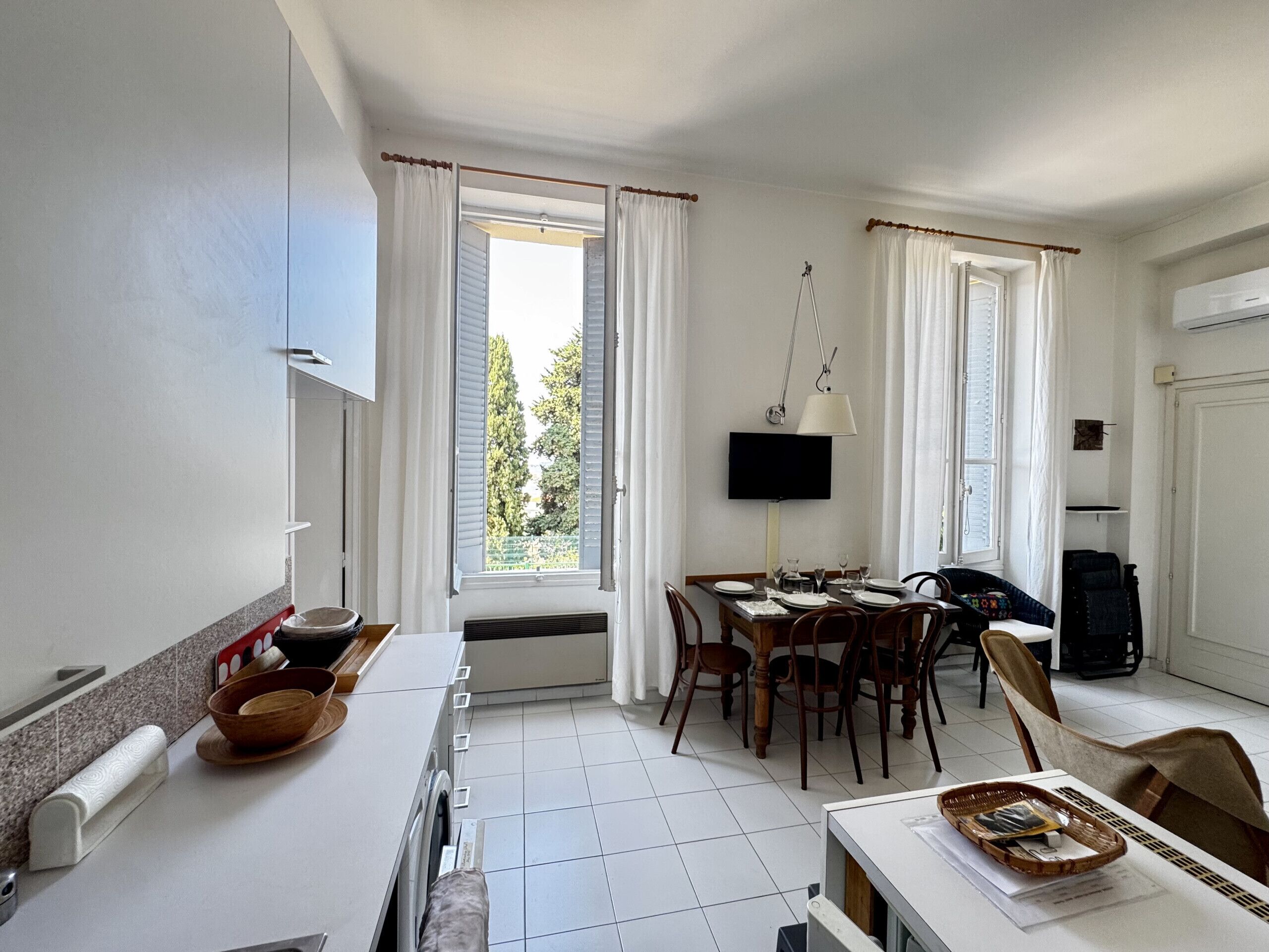 A hotel room with a dining area, a TV mounted on the wall, and a window with curtains.