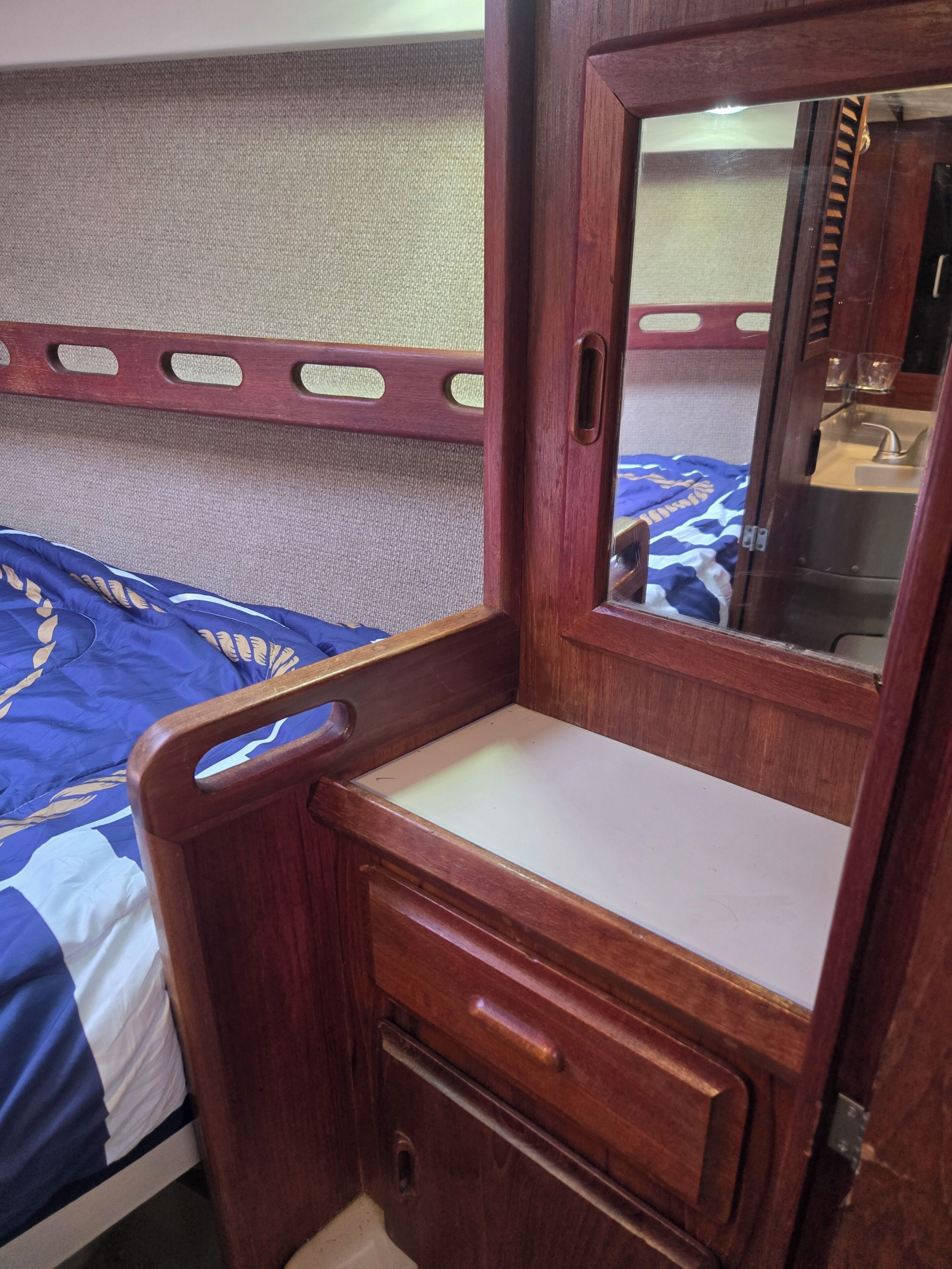 A wooden cabinet with a mirror and a small drawer, situated next to a bed with a blue and white patterned bedspread.