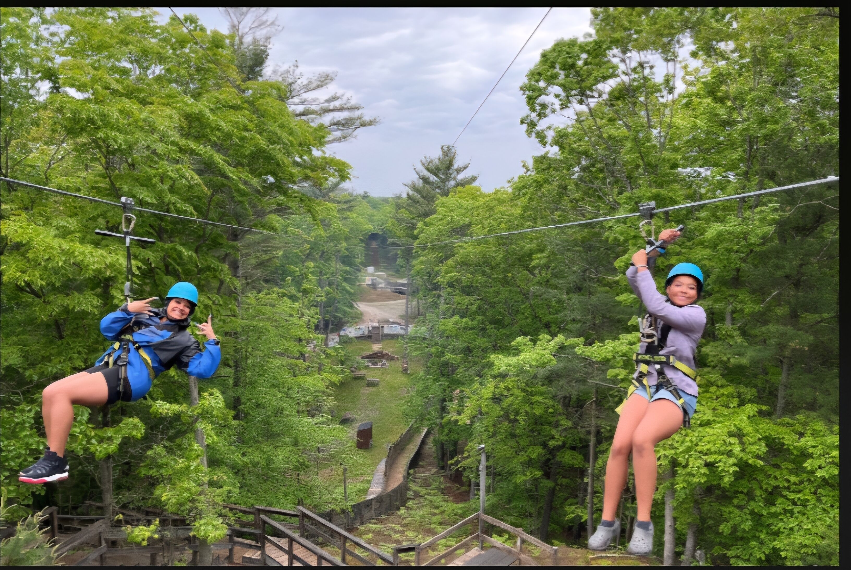 Two individuals zip-lining through a forested area with wooden walkways and a distant view of a building.