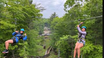 Two individuals zip-lining through a forested area with wooden walkways and a distant view of a building.