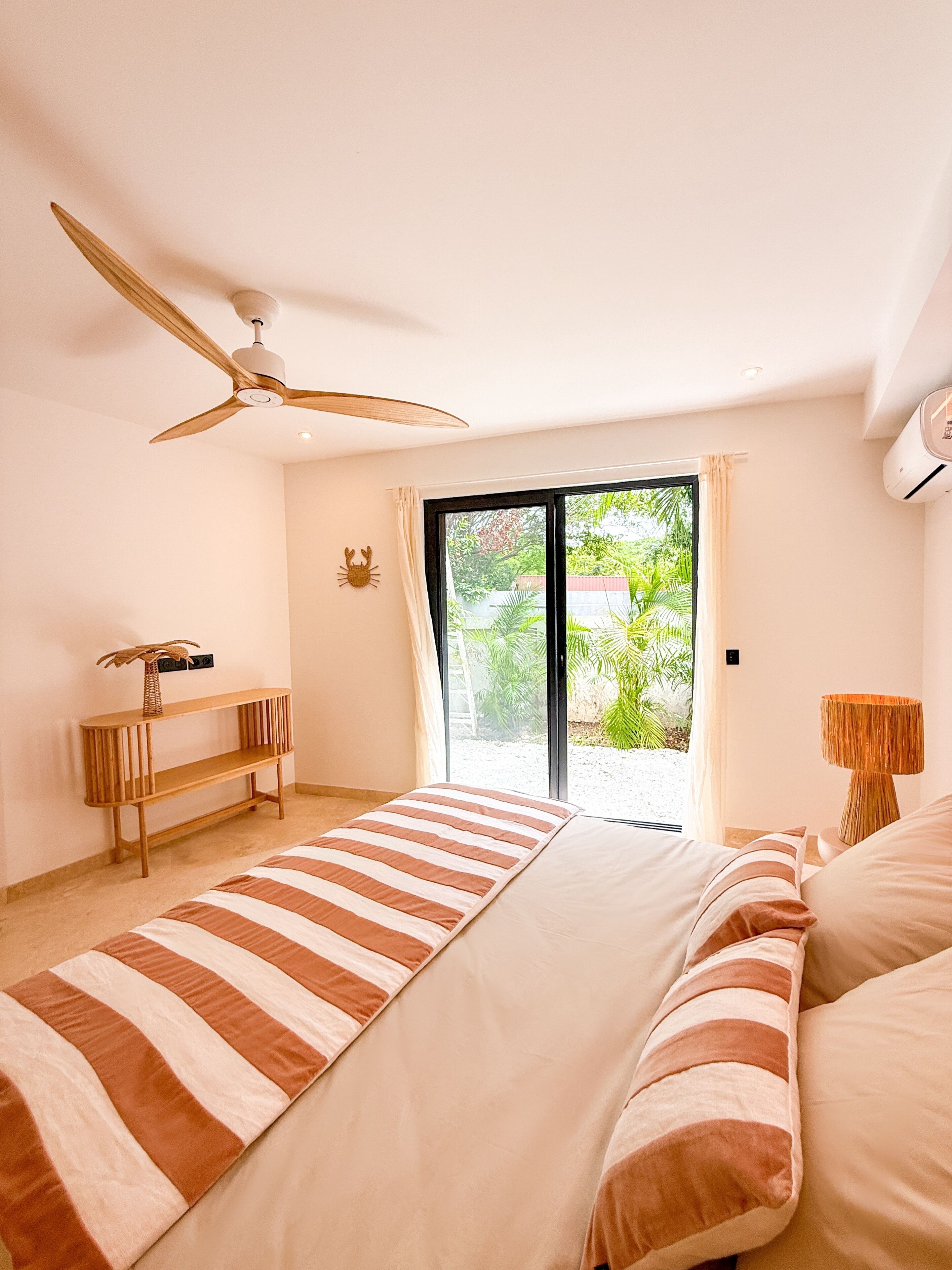 A bedroom with a bed, a ceiling fan, a window with a view of greenery, a bedside table, and a lamp.