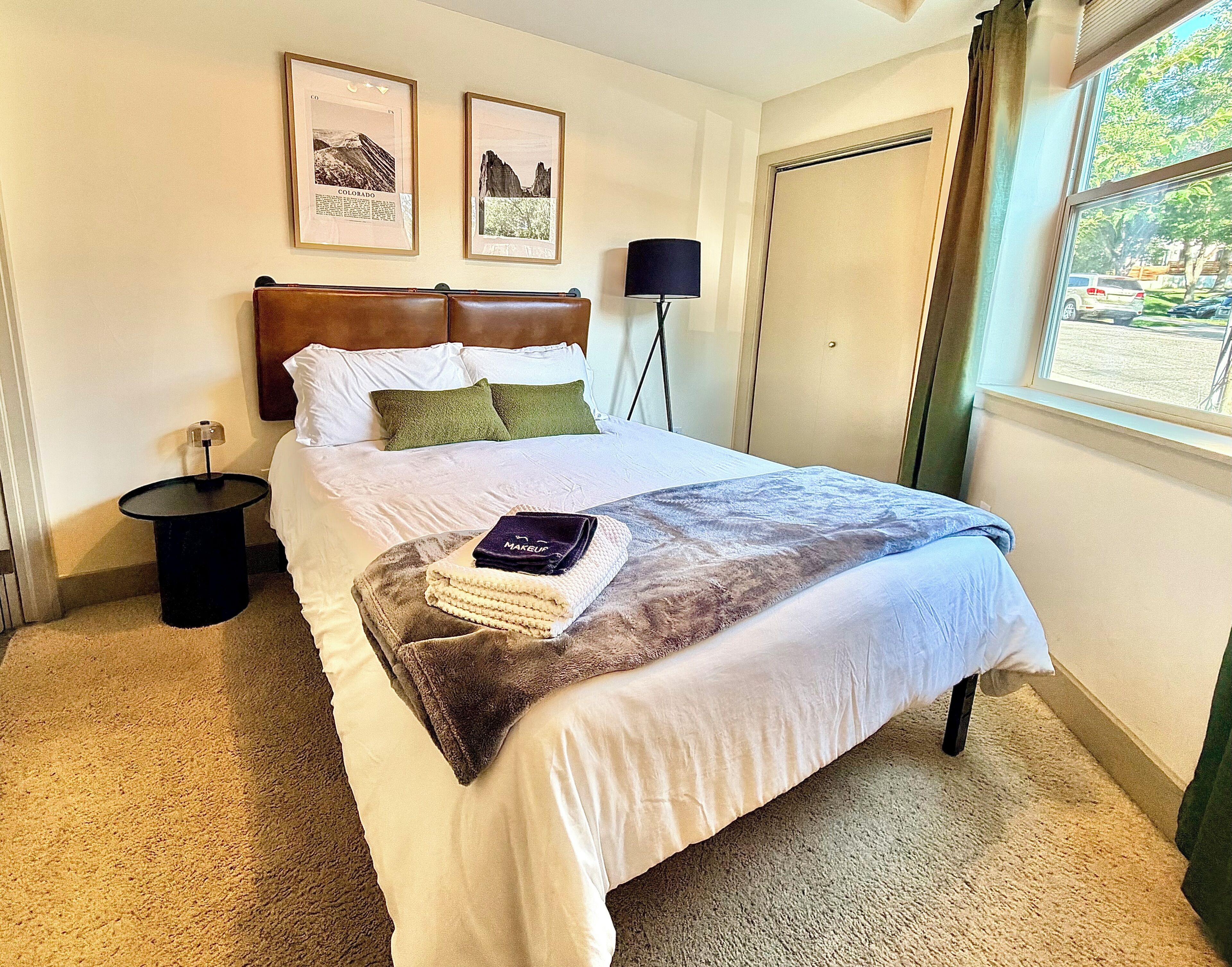 A hotel room with a bed, a nightstand, a lamp, a window with a view, and two framed pictures on the wall.